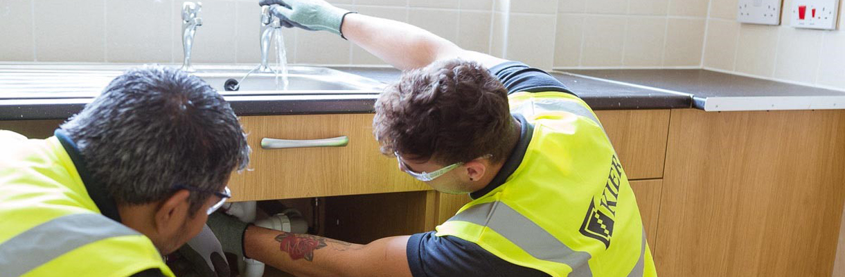 Image of two Kier employees undertaking some plumbing works with one adjusting the pipes under the sink while turning on the tap